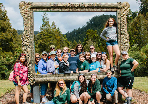 Group of students posing inside a large decorative frame at a scenic park.