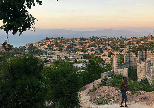 View of a hillside town at sunset with a person walking along a dirt path.