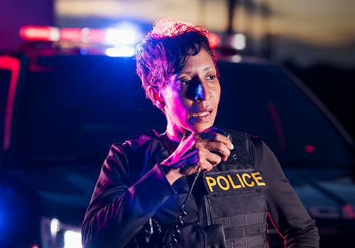 Police officer speaking into a radio in front of a patrol vehicle with flashing lights