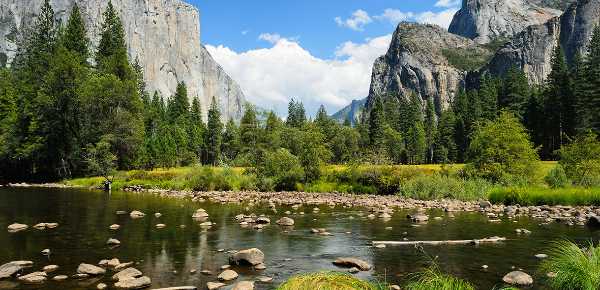 River flowing through Yosemite Valley with granite cliffs and pine trees under a blue sky.